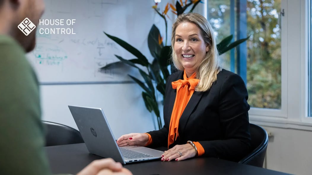 A blonde woman in an orange blouse and a black blazer is in a conversation with her colleague. 