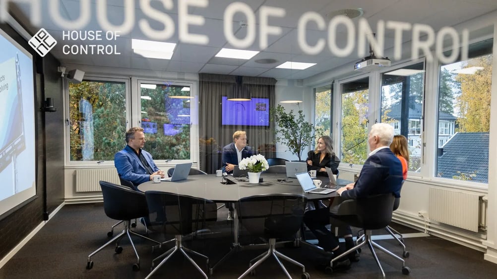 A group of colleagues in a meeting room having a discussion around a large table. House of Control logo visible on the glass wall.