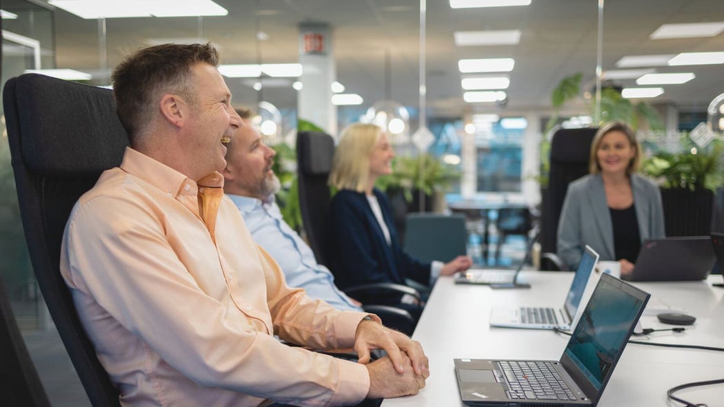 A group of people sitting down at a desk in an office enviroment with their laptops on the desk. They seem to conversate happily together.
