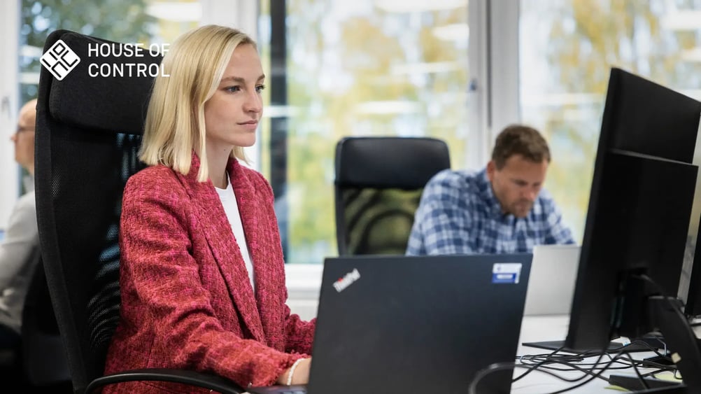 A woman in a red blazer is working and looking at a monitor. A man in a checkered shirt is sitting next to her in the background working on a laptop. House of Control logo in the upper left corner.