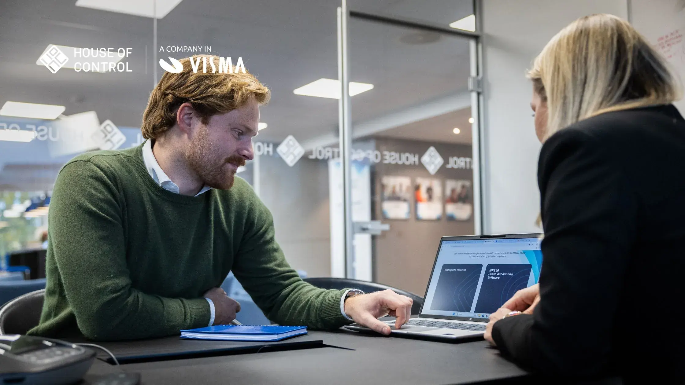 A man in a green shirt is showing a blonde woman something on the PC at the office