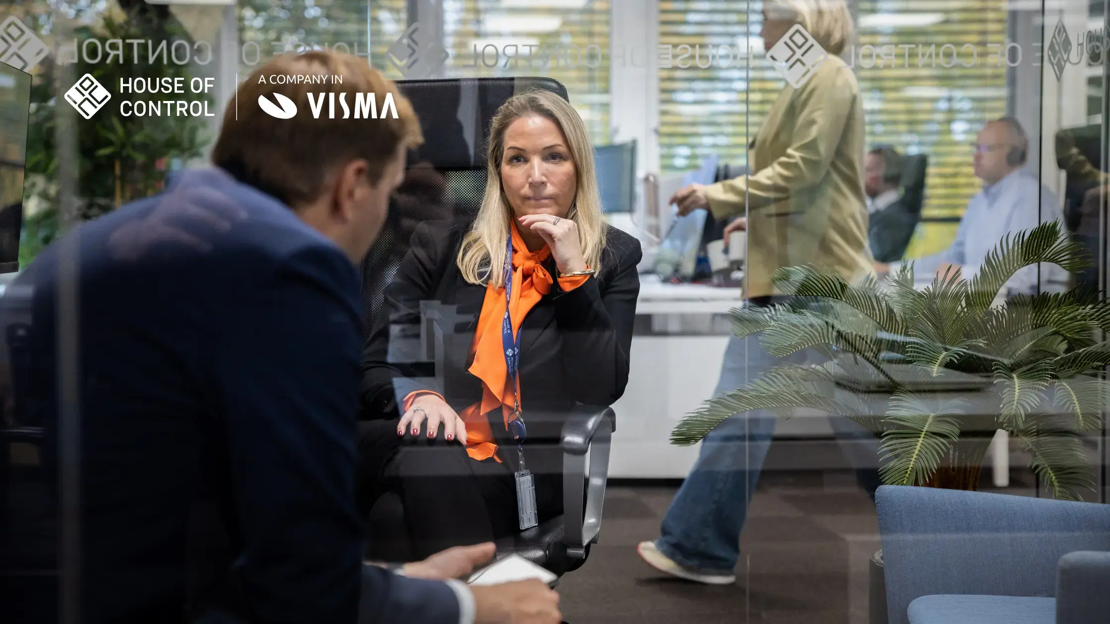 A blonde woman in an orange blouse and a black blazer is in a conversation with her colleague. 