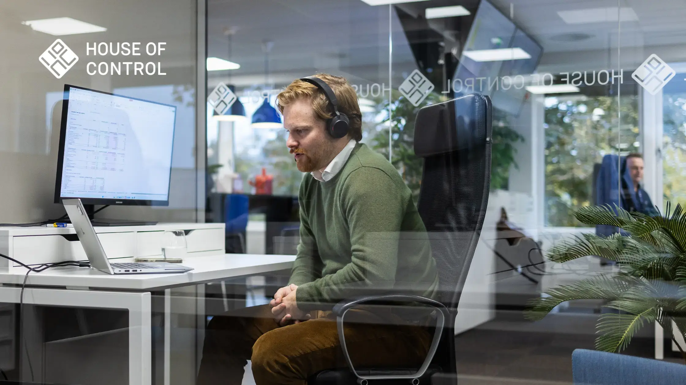 A man in a green sweater is sitting in front of the computer with a headset in a meeting room