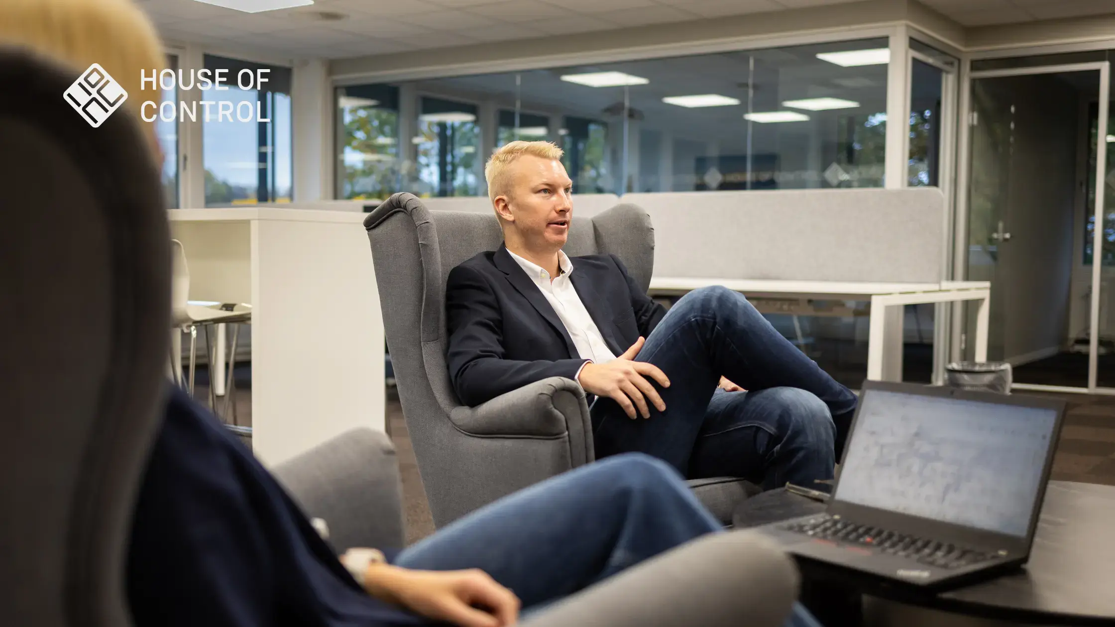A blonde man is sitting with his legs crossed in a chair, looking like he's in a conversation at the office