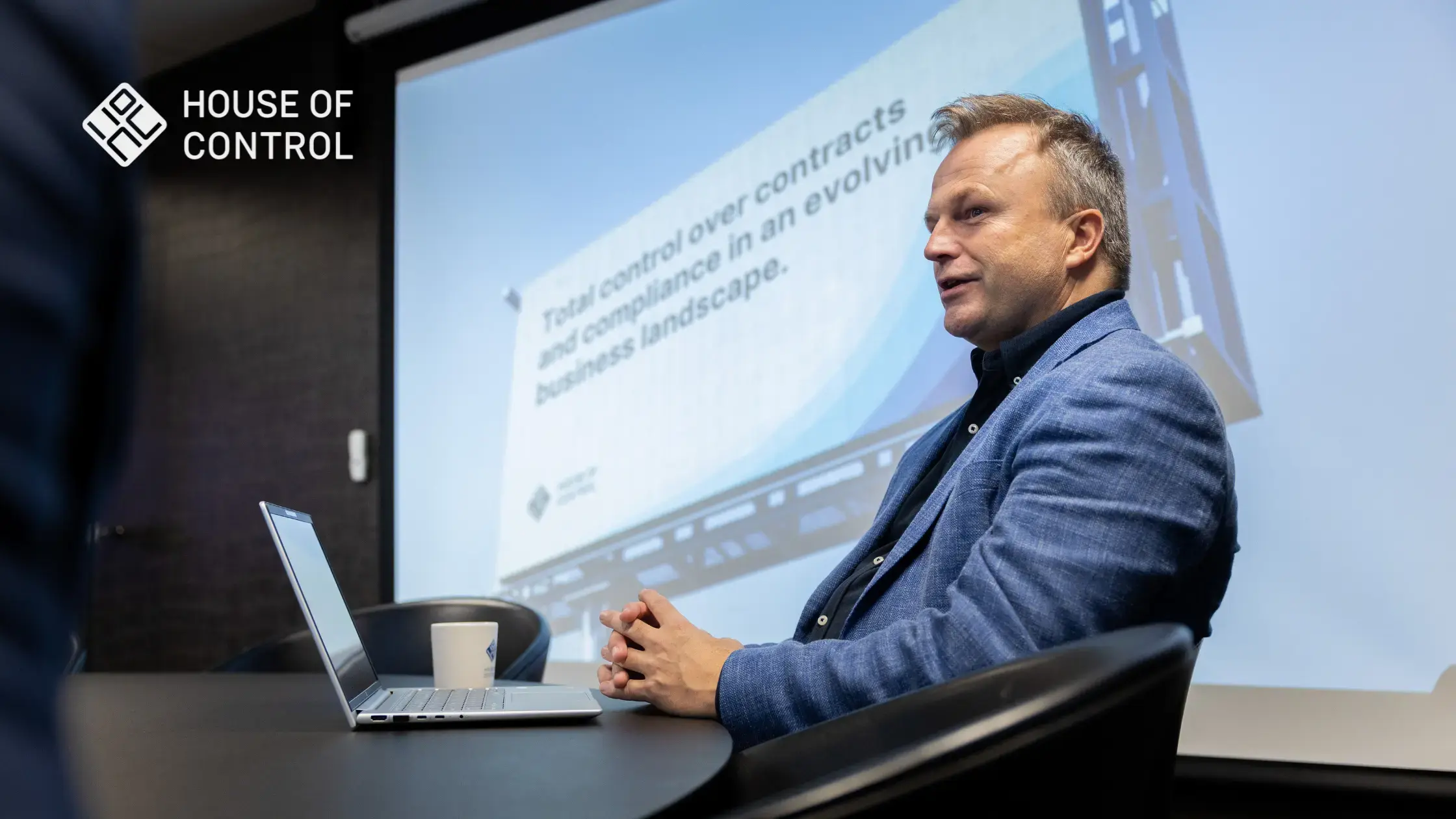 A House of Control employee sitting down in a meeting with a big screen behind him