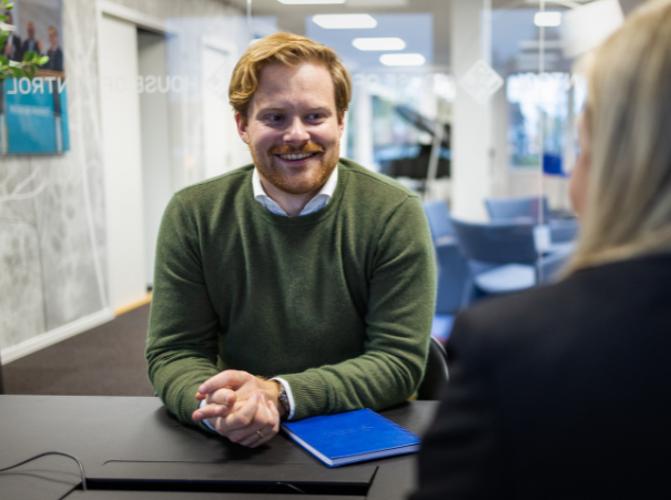 The image shows a person with a blurred face sitting at a desk in an office environment, facing another individual whose back is to the camera. The person at the desk is wearing a green sweater and has their hands clasped together. In front of them is a blue notebook or folder. The background suggests a modern office space with a poster on the wall and a bright, natural light source from the ceiling.