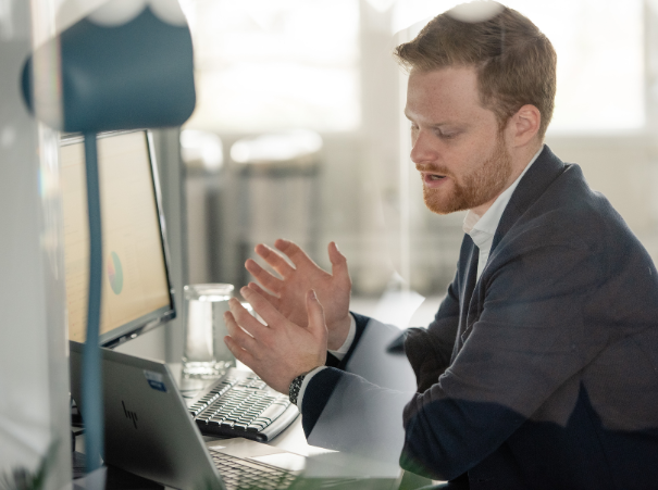 The image depicts a professional setting where a man appears to be engaged in a discussion or a meeting. He is seated at a desk, wearing a suit and tie, and has his hands clasped together in front of him, possibly indicating active engagement or concentration on a matter of discussion. On his desk, there is a laptop, suggesting that he may be involved in work or communication that requires technology. The office environment is modern and bright, with large windows allowing natural light to fill the space. There are no visible texts or other individuals in the image.