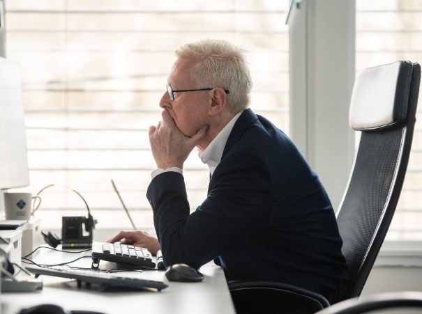 The image shows an older adult sitting at a desk in what appears to be an office environment. The person is wearing a dark blazer and is seated on a black office chair. The desk is equipped with a computer setup that includes a monitor, a keyboard, and a mouse, as well as other items that suggest a workspace, such as papers and possibly documents or books. The individual seems to be in thought, with their hand on their chin. The room has a window with blinds, allowing natural light to enter, and the overall setting is professional and tidy.