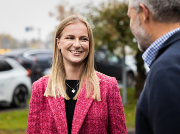 The image shows a woman in a bright pink tweed blazer and a black top, smiling and talking to a man in a blue sweater. They appear to be outdoors with a blurred background that includes greenery and a white car.