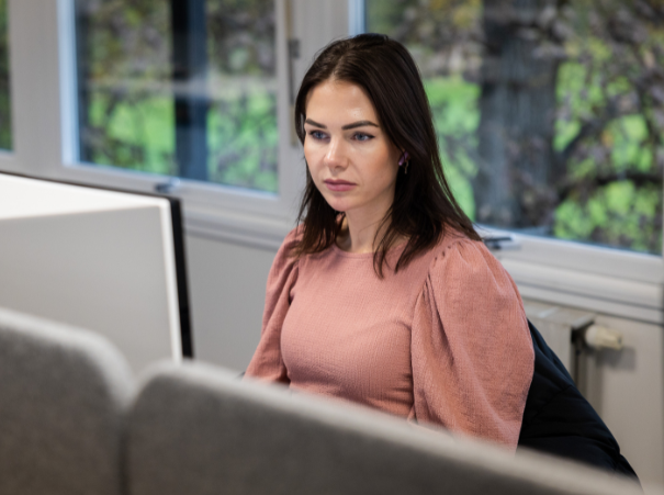The image shows a person sitting in a room with a window behind them that overlooks a green outdoor area. The person is wearing a pink top and is seated in front of a computer monitor with a grey frame. The lighting in the room is bright, suggesting it is daytime.