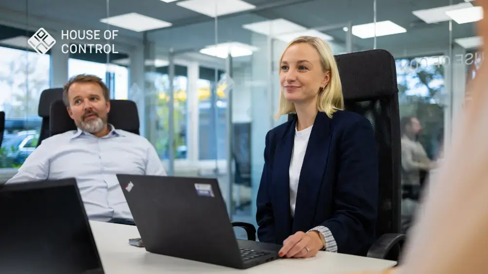 An office setting with a man and a woman seated at a table with laptops open in front of them. They are in a meeting and looking towards someone off-camera. A logo in the top left corner that reads 'HOUSE OF CONTROL.'