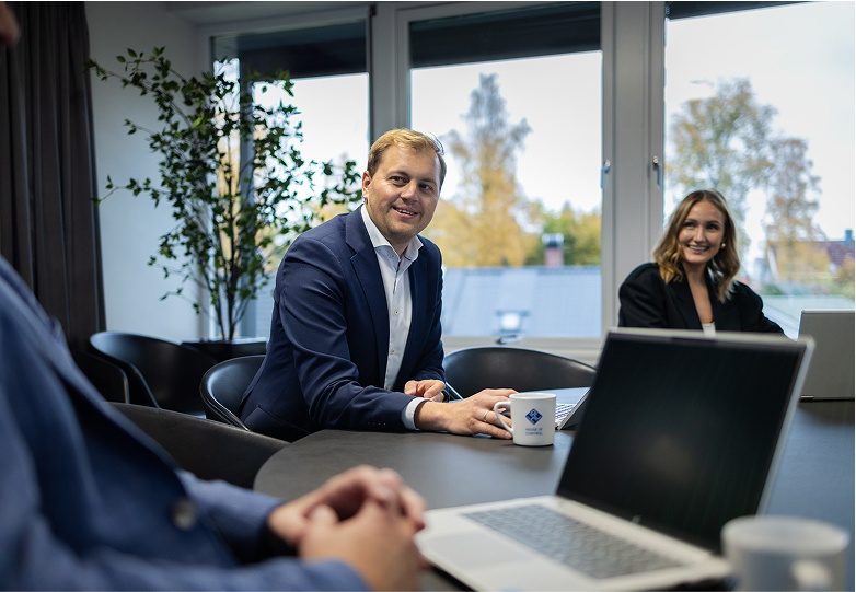 A group of professionally dressed colleagues sit together in a bright meeting room, smiling and engaged in a lively discussion about DORA compliance. A laptop rests on the desk in front of them, with large windows in the background letting in natural light.