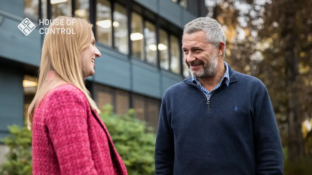 To personer står utendørs og smiler til hverandre, med en kontorbygning i bakgrunnen. En vennlig og profesjonell atmosfære i et forretningsmiljø.