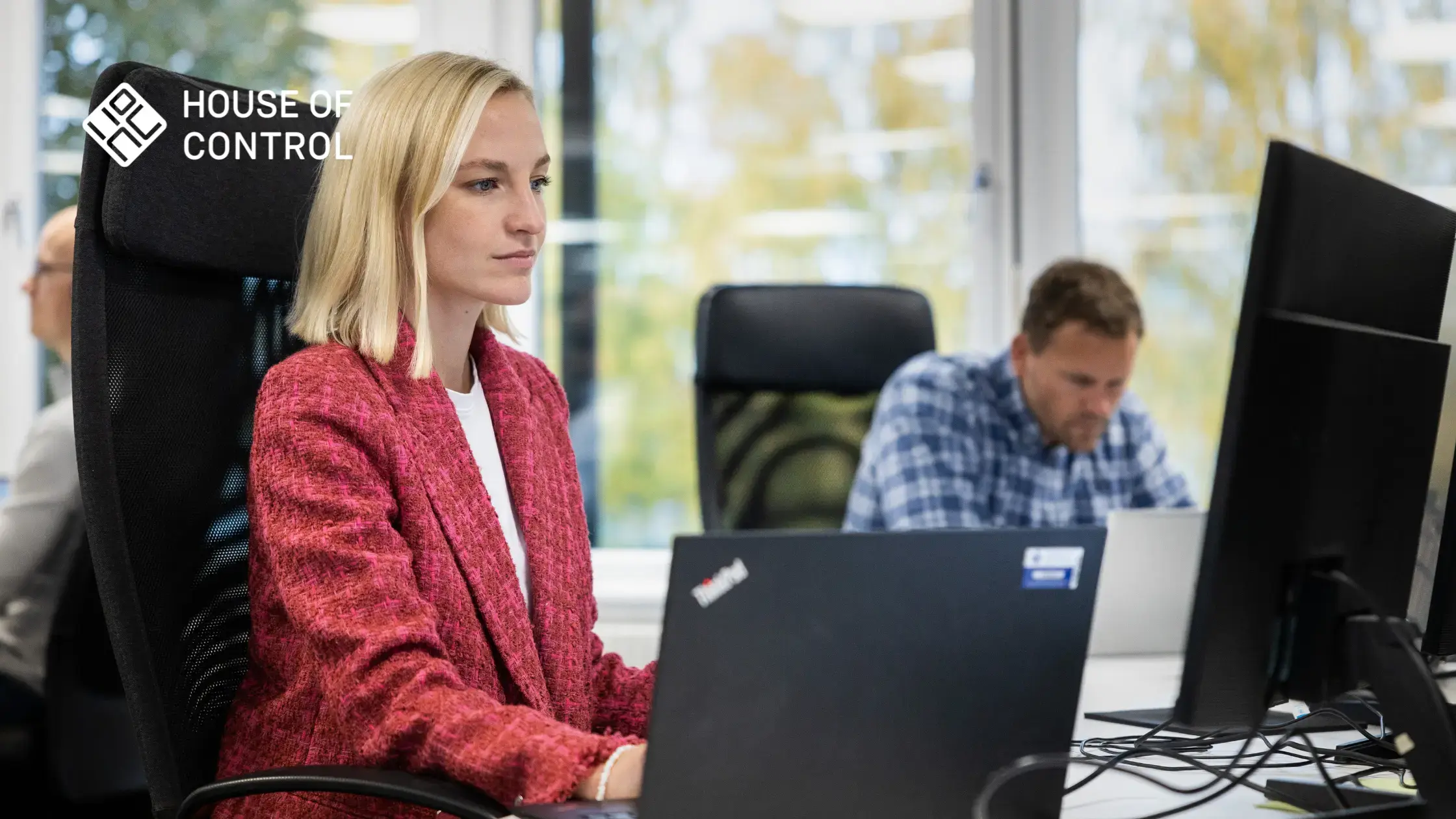 A woman in a red blazer is working and looking at a monitor. A man in a checkered shirt is sitting next to her in the background working on a laptop. House of Control logo in the upper left corner.